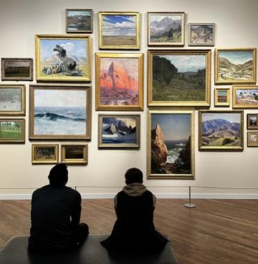 Two people sit on a bench admiring framed scenes of nature on a gallery wall